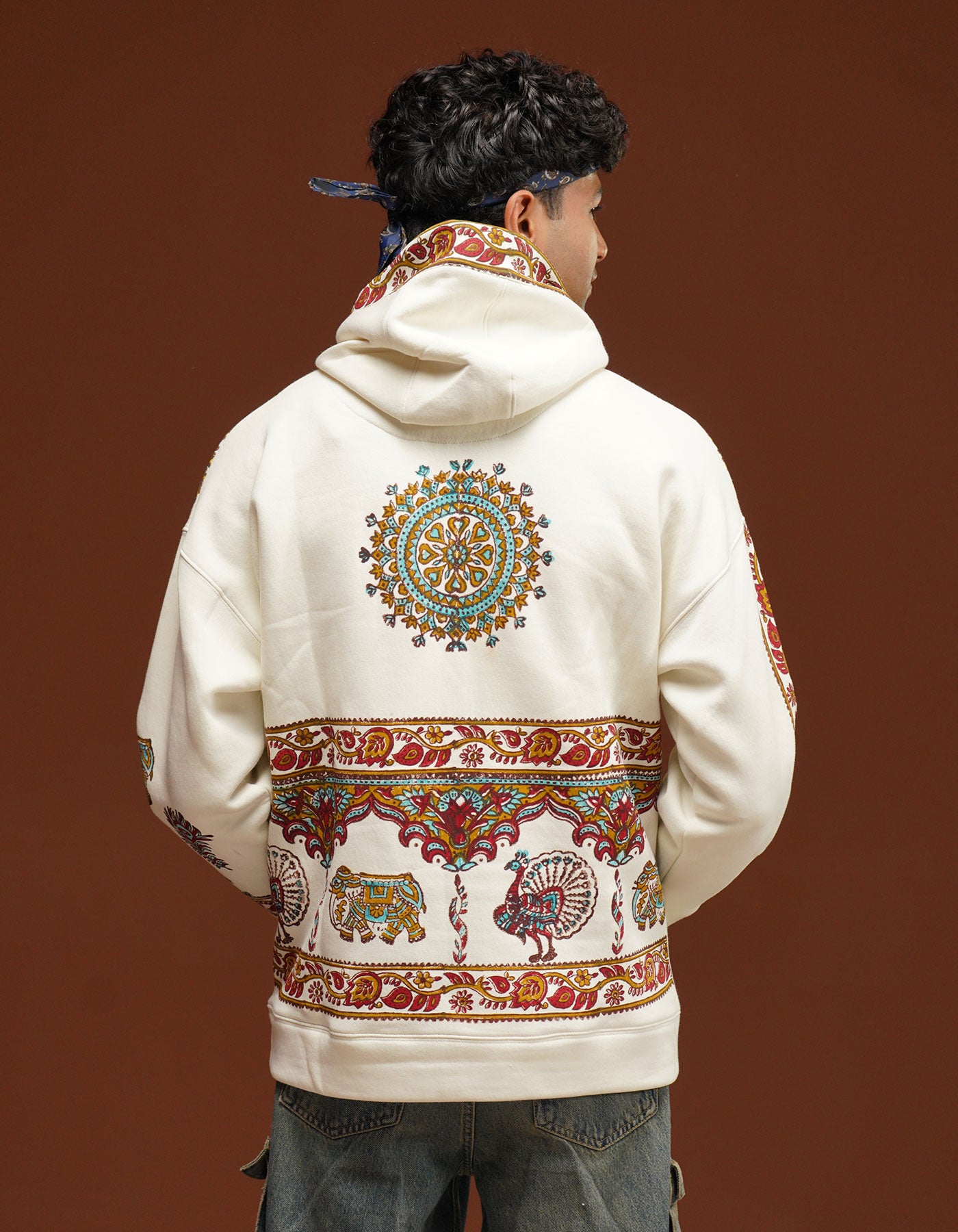 Person wearing a cream-colored hoodie with intricate patterns on the back against a brown background