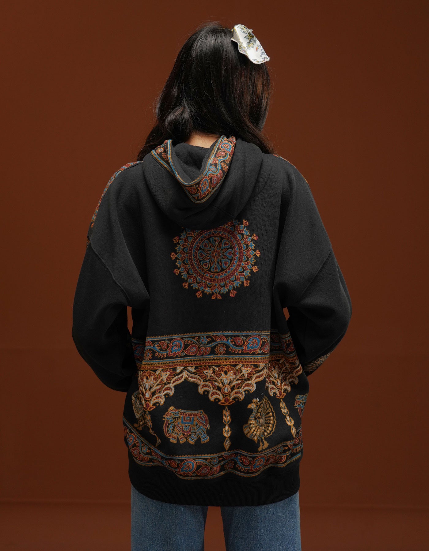 Person wearing a black hoodie with intricate patterns on the back against a brown background