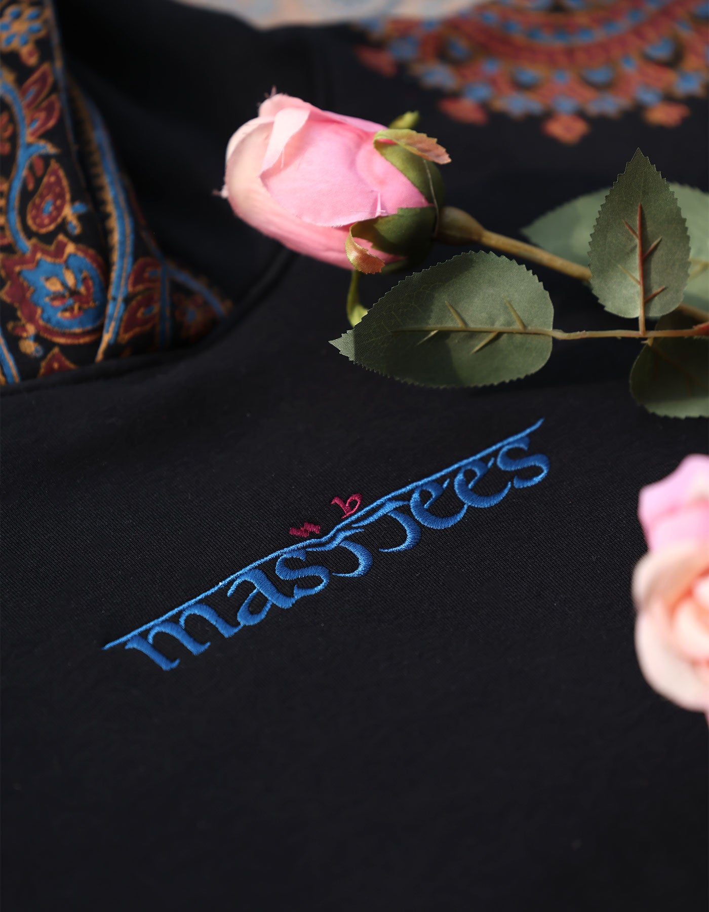 Black garment with 'massy fees' embroidered in blue, surrounded by pink roses and green leaves.