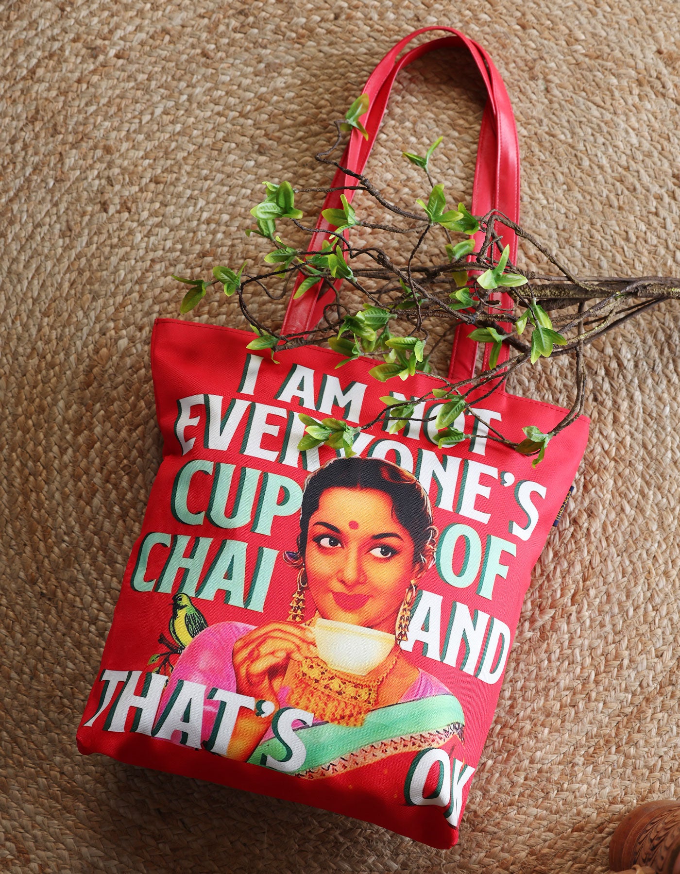 Red tote bag with a graphic of a woman holding a cup and text, hanging on a textured wall.