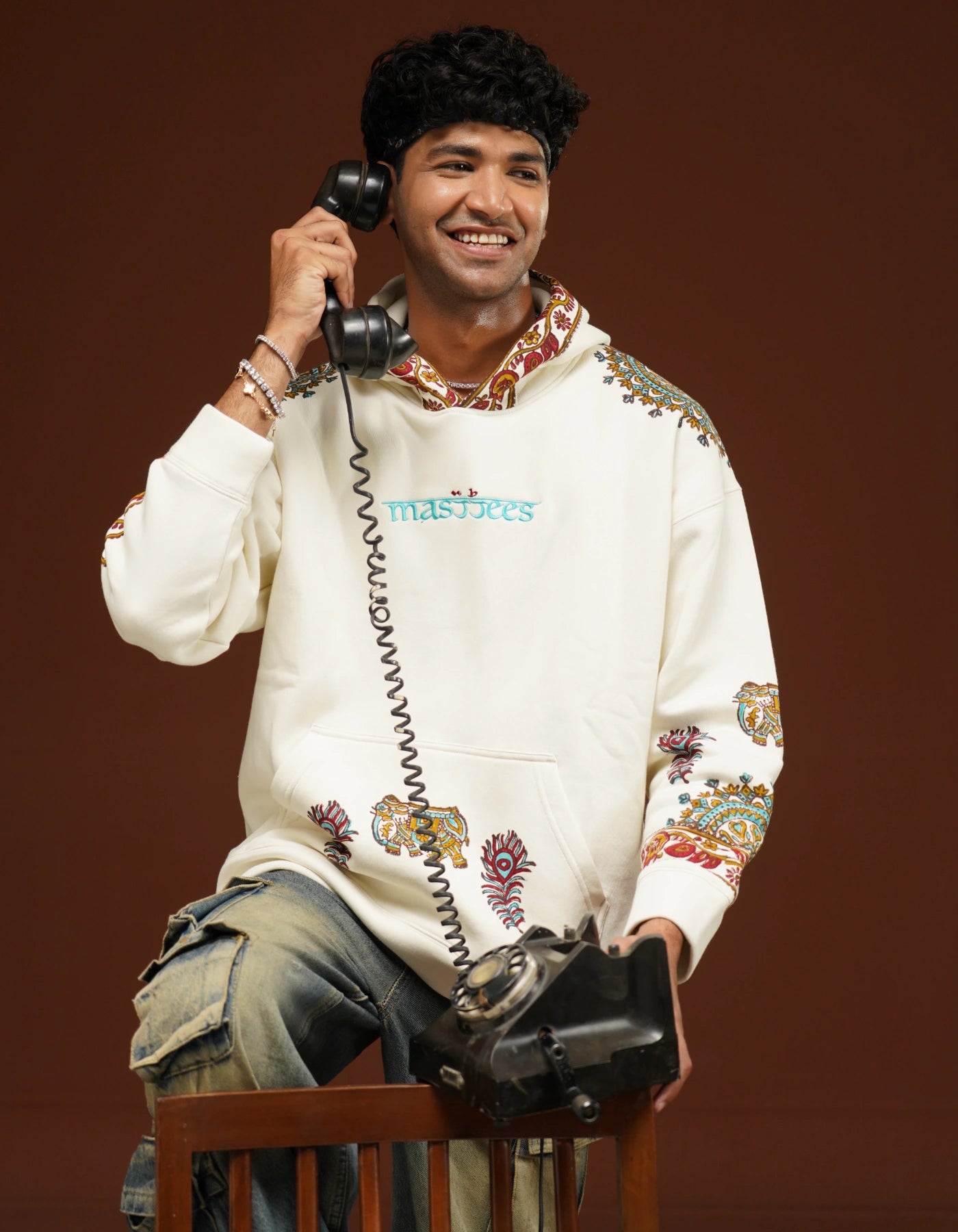 Person wearing a white hoodie with colorful patterns, sitting on a chair and holding a vintage phone.