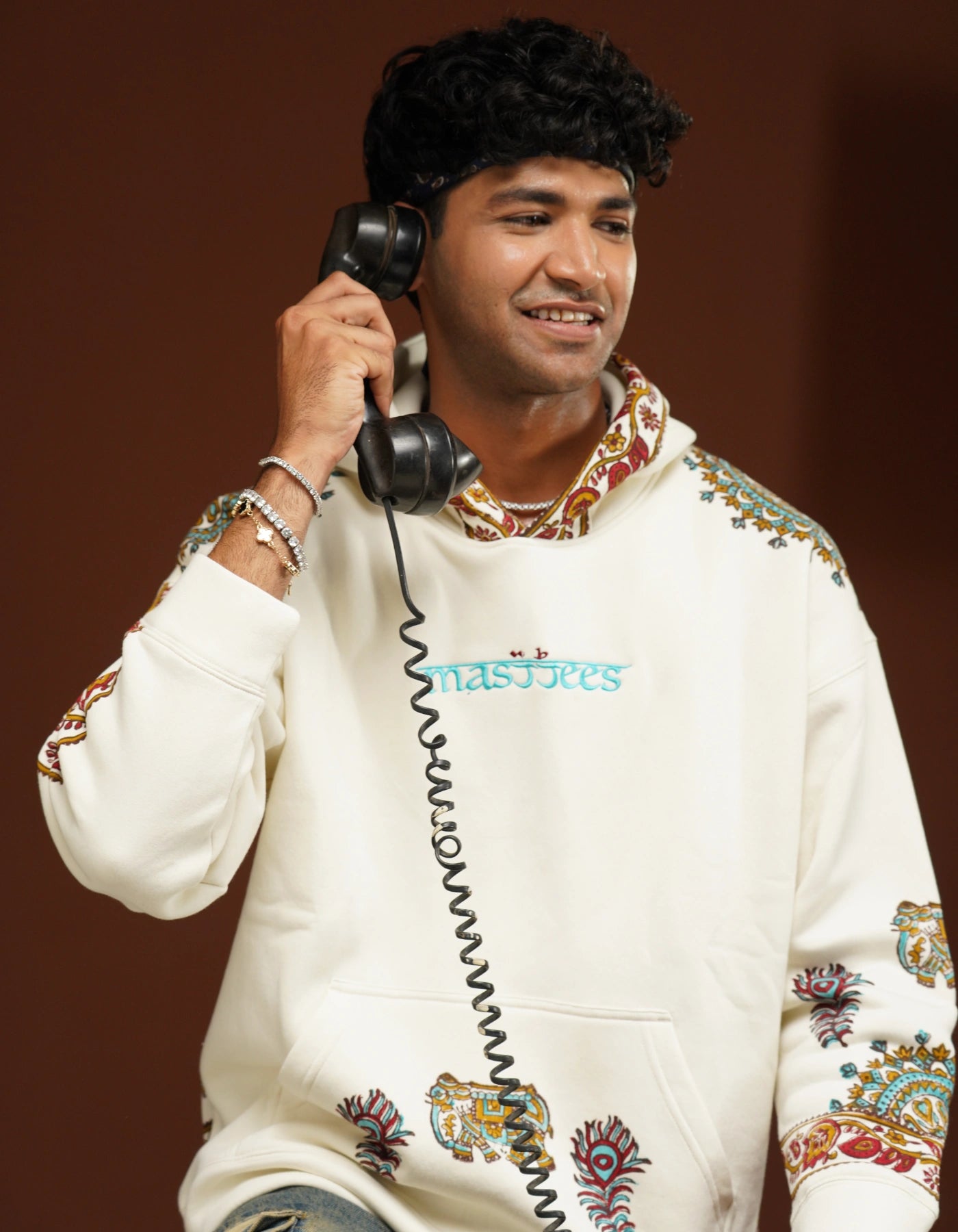 Person wearing a white hoodie with colorful patterns and text, holding a black phone receiver against a brown background.