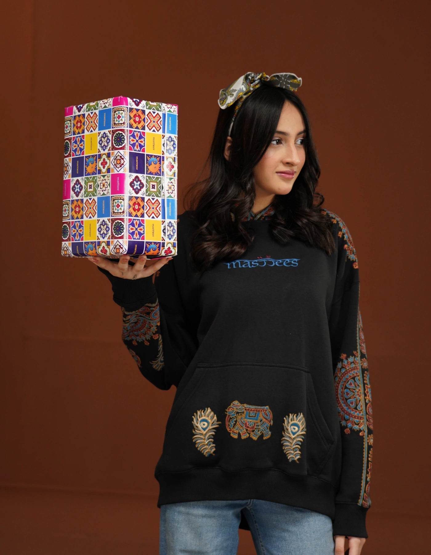 Woman holding a colorful box with a pattern, wearing a black hoodie with embroidered designs on a brown background.